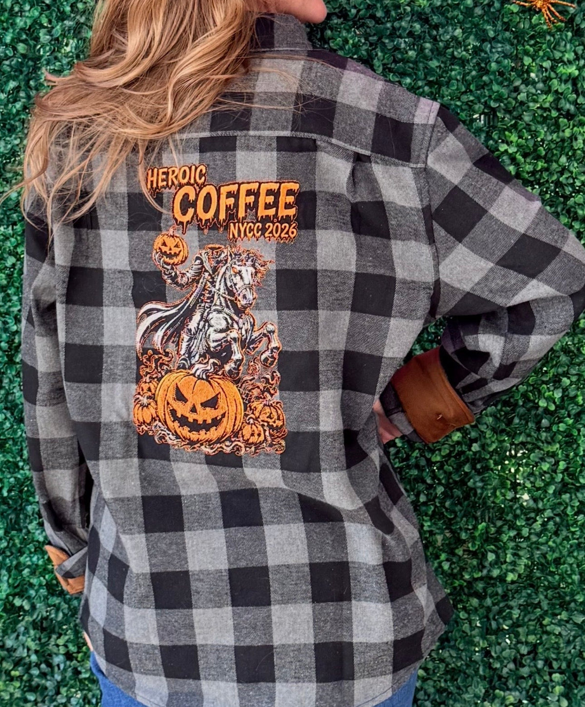 A woman shown from the back wearing a black and gray buffalo plaid flannel shirt with a large orange and white graphic on the back that reads 'HEROIC COFFEE NYC 2026' and features a skeleton and jack-o'-lantern. She is standing in front of a green artificial hedge wall decorated with Halloween props.